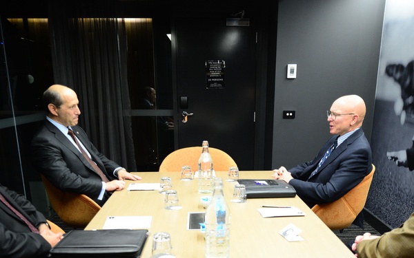 Four men in formal business attire are seated around a rectangular conference table in a modern meeting room, engaged in what appears to be a formal bilateral or diplomatic discussion. The room features dark walls with what appears to be a military aircraft mural on the right side, orange upholstered chairs, and a sign on the door indicating room capacity. Documents, water bottles, glasses, and a laptop are visible on the table.
