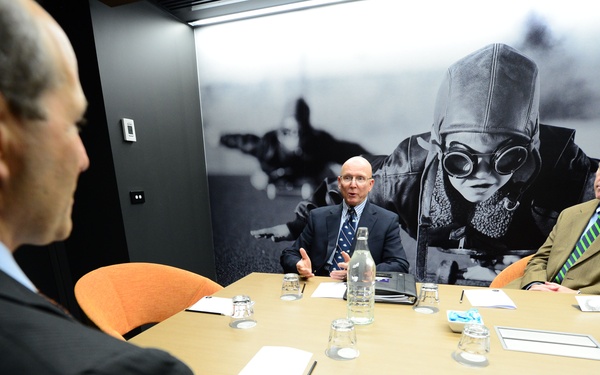 A formal meeting is taking place in a modern conference room between three men in business attire seated around a light-colored table. The room features a large black-and-white photographic mural on the back wall depicting a person wearing goggles and a helmet. The setting appears professional, with notepads, water bottles, and glasses arranged on the table.