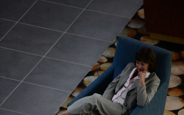 An overhead view of two professionally dressed individuals seated in blue upholstered armchairs in what appears to be a hotel or corporate lobby. A woman in a gray blazer and a man in a dark suit appear to be engaged in conversation. The setting features a distinctive patterned carpet with cross-sections of wood log designs.