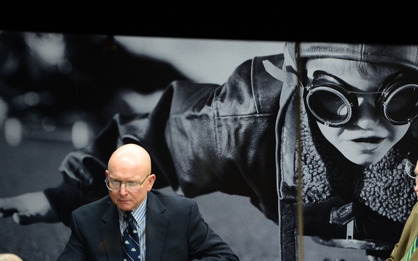 A formal meeting or briefing is taking place around a conference table with at least four men in business attire. The setting features a large black-and-white photographic mural on the wall depicting what appears to be a World War II-era aviator or pilot wearing goggles and a leather flight jacket. The scene is photographed through glass, creating a slightly reflective framing effect.