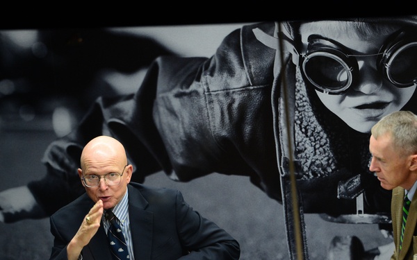 A formal meeting or panel discussion is taking place around a conference table, with several individuals seated and engaged in conversation. A bald man in a navy suit appears to be speaking or making a point, while a man in a tan suit listens attentively. Behind them is a large black-and-white photographic backdrop depicting a child wearing aviator goggles and a leather jacket.