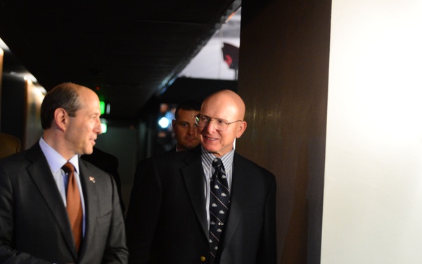 Four men in business attire are walking together through a dimly lit corridor or hallway. The two men in the foreground appear to be engaged in conversation while walking. The setting appears to be an indoor venue with modern architectural elements.