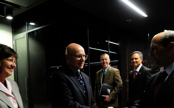 A formal greeting scene in what appears to be a modern building lobby or corridor. Two men in dark suits are shaking hands in the foreground, while a woman in a gray blazer stands nearby smiling, and two additional men in suits observe in the background. The setting suggests an official or professional reception.