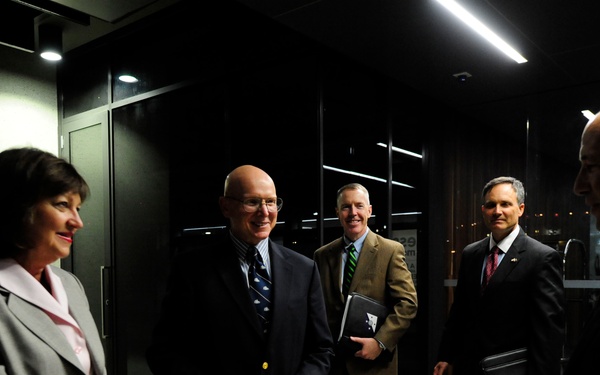 Five formally dressed individuals are gathered in what appears to be a modern building lobby or corridor, engaged in conversation. The group consists of four men in business suits and one woman in a gray blazer, suggesting a professional or official meeting. The setting features dark architectural elements with overhead fluorescent lighting.