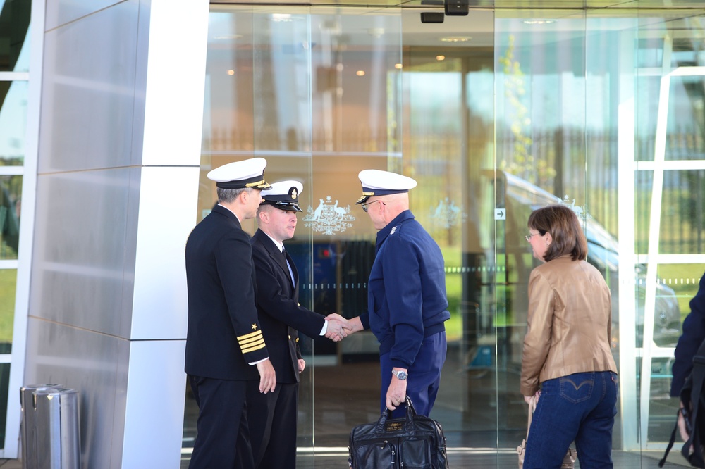 The image shows a formal greeting taking place outside what appears to be an official government building, identifiable by the Australian Commonwealth Coat of Arms visible on the glass doors. Two naval officers in dark uniforms with white-topped caps are shaking hands with a third uniformed individual in a blue jacket and white cap. A civilian woman in a tan jacket and jeans stands nearby, suggesting this is an official reception or arrival ceremony.