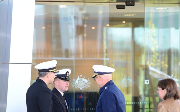 The image shows a formal greeting taking place outside what appears to be an official government building, identifiable by the Australian Commonwealth Coat of Arms visible on the glass doors. Two naval officers in dark uniforms with white-topped caps are shaking hands with a third uniformed individual in a blue jacket and white cap. A civilian woman in a tan jacket and jeans stands nearby, suggesting this is an official reception or arrival ceremony.