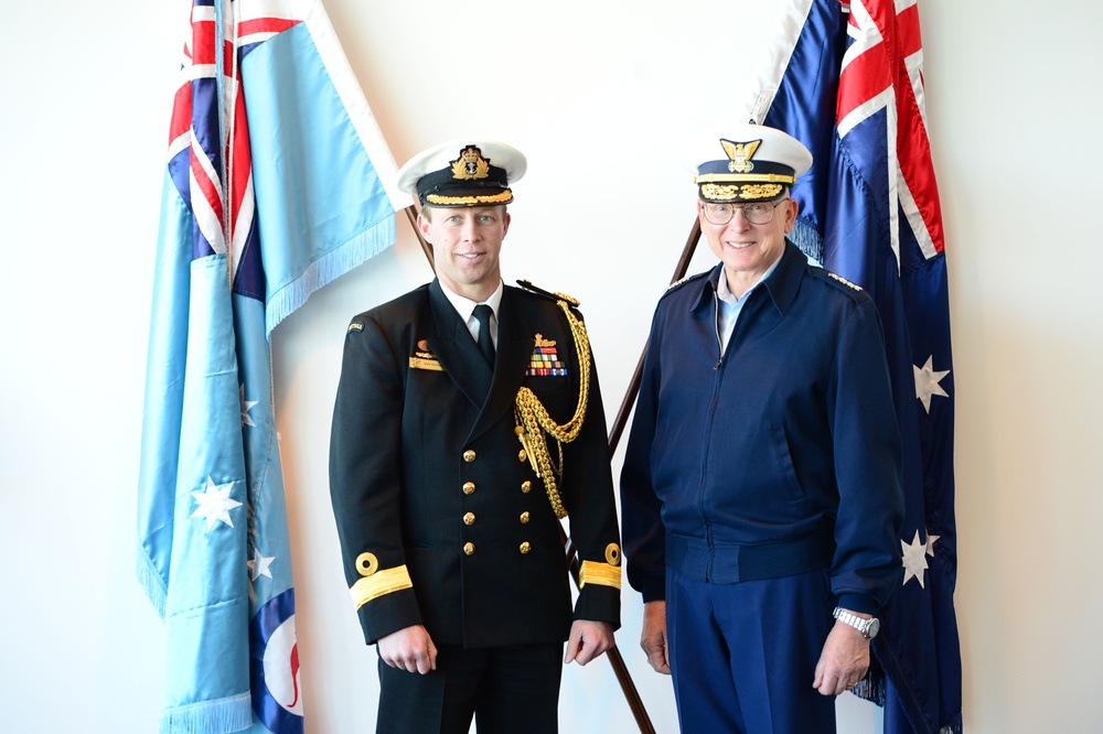 Two military officers in formal dress uniforms pose together in front of flags. The officer on the left wears a dark navy ceremonial uniform with gold aiguillettes, medal ribbons, and a white-topped peaked cap, consistent with a senior Royal Australian Navy officer. The officer on the right wears a blue service jacket and white-topped peaked cap with gold insignia, consistent with a senior US Coast Guard officer.
