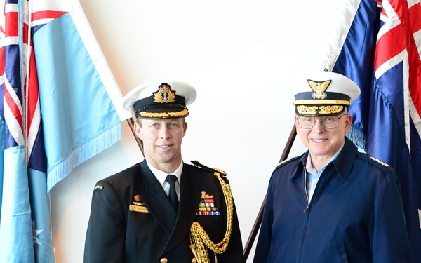 Two military officers in formal dress uniforms pose together in front of flags. The officer on the left wears a dark navy ceremonial uniform with gold aiguillettes, medal ribbons, and a white-topped peaked cap, consistent with a senior Royal Australian Navy officer. The officer on the right wears a blue service jacket and white-topped peaked cap with gold insignia, consistent with a senior US Coast Guard officer.