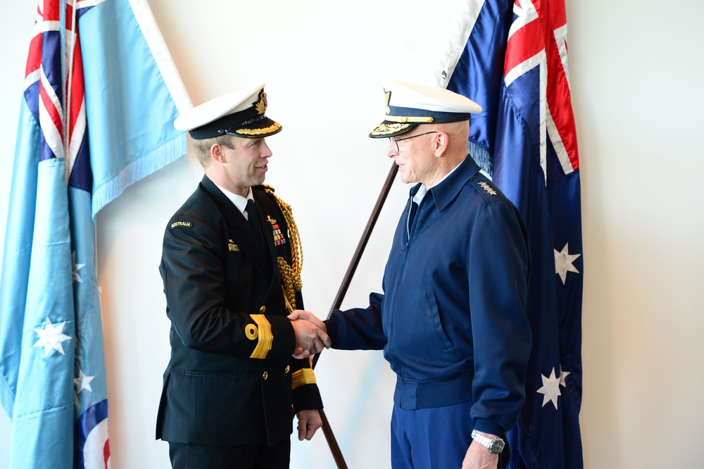 Two senior military officers are shaking hands in what appears to be a formal ceremony or official meeting. The officer on the left wears an Australian Navy dress uniform with gold aiguillettes and an 'AUSTRALIA' name tape, while the officer on the right wears a dark blue military jacket with four-star rank insignia. Both wear peaked caps with gold insignia, and one officer holds a ceremonial cane or baton.