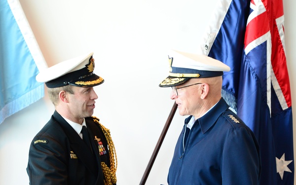 Two senior military officers are shaking hands in what appears to be a formal ceremony or official meeting. The officer on the left wears an Australian Navy dress uniform with gold aiguillettes and an 'AUSTRALIA' name tape, while the officer on the right wears a dark blue military jacket with four-star rank insignia. Both wear peaked caps with gold insignia, and one officer holds a ceremonial cane or baton.