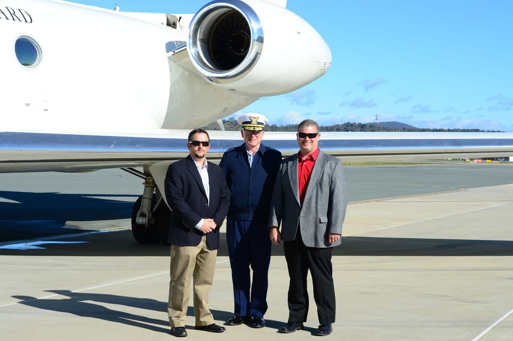Three men are posing for a photograph on an airport tarmac in front of a white private/executive jet. The central figure is wearing a military or naval dress uniform with a white peaked cap, flanked by two men in civilian business attire. The setting appears to be a sunny day at a regional airport.