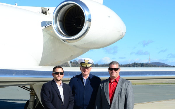 Three men are posing for a photograph on an airport tarmac in front of a white private/executive jet. The central figure is wearing a military or naval dress uniform with a white peaked cap, flanked by two men in civilian business attire. The setting appears to be a sunny day at a regional airport.