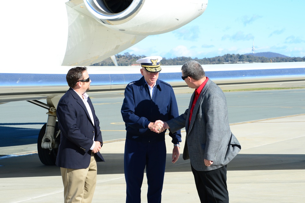Three men are standing on an airport tarmac beside a white aircraft. A uniformed military or coast guard officer in a dark blue uniform and white peaked cap is shaking hands with a man in a gray jacket and red shirt, while a third man in a navy blazer and khaki pants observes. The setting appears to be an official arrival or greeting ceremony at an airfield.