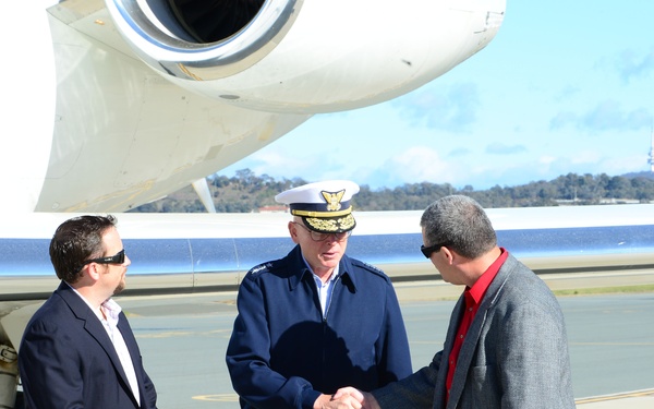 Three men are standing on an airport tarmac beside a white aircraft. A uniformed military or coast guard officer in a dark blue uniform and white peaked cap is shaking hands with a man in a gray jacket and red shirt, while a third man in a navy blazer and khaki pants observes. The setting appears to be an official arrival or greeting ceremony at an airfield.