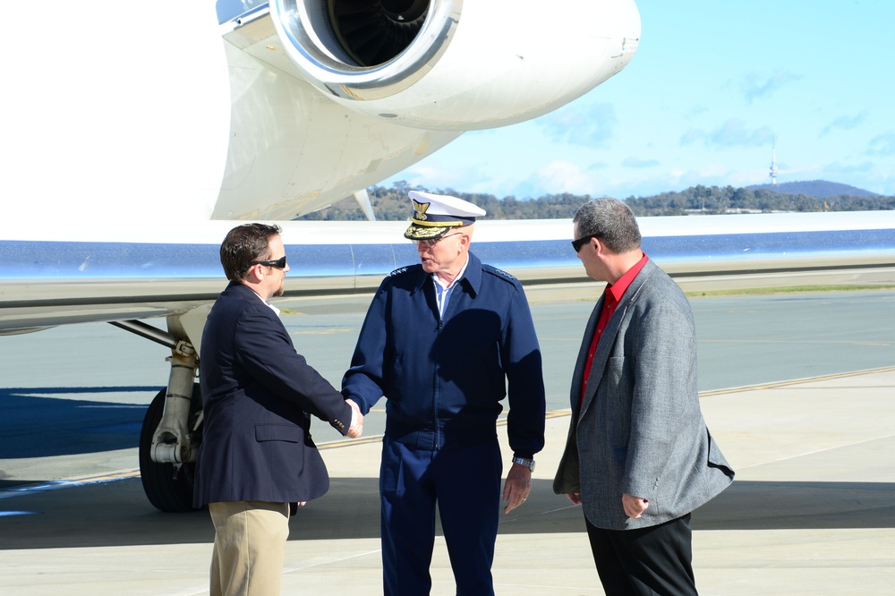 Three men are standing on an airport tarmac beside a white jet aircraft. A uniformed military officer wearing a white peaked cap and navy blue uniform is shaking hands with a man in a dark suit, while a third man in a grey jacket and red shirt stands nearby observing. The setting appears to be a formal arrival or greeting ceremony at an airfield.