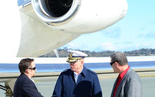 Three men are standing on an airport tarmac beside a white jet aircraft. A uniformed military officer wearing a white peaked cap and navy blue uniform is shaking hands with a man in a dark suit, while a third man in a grey jacket and red shirt stands nearby observing. The setting appears to be a formal arrival or greeting ceremony at an airfield.