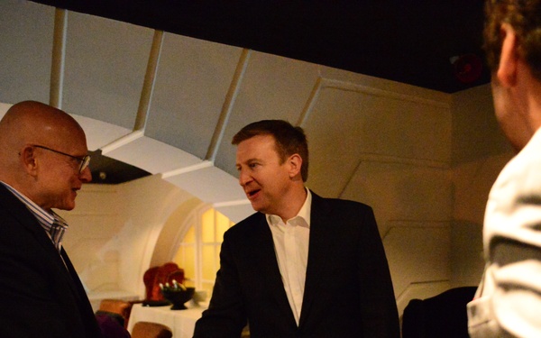 Two men in dark suits are shaking hands in what appears to be an upscale restaurant or event venue with distinctive arched white ceiling panels. The man on the left is bald and wearing glasses, while the man on the right is taller with light brown hair. A third person in a light-colored jacket is partially visible in the foreground right.