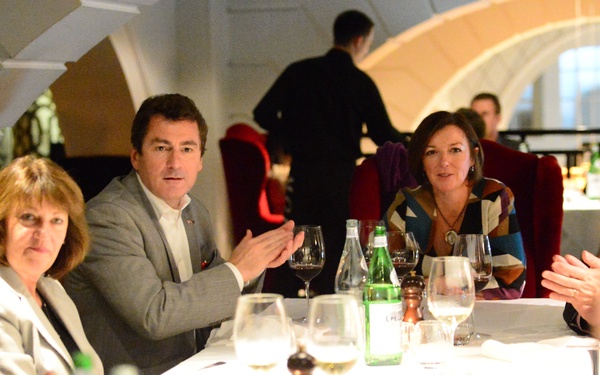 A group of professionals are gathered around a white-clothed dining table in an upscale restaurant setting. The scene appears to be a business dinner or formal social gathering, with multiple people engaged in conversation. The restaurant features distinctive arched architectural elements in the background.