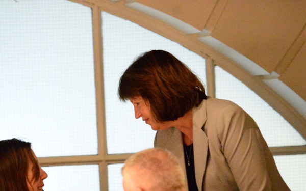 The image shows an indoor dining or event setting where a standing woman in a beige blazer is leaning down to speak with a seated woman who is smiling. Several other attendees are visible in the foreground and background. The setting appears to be a formal or semi-formal gathering, possibly a dinner event or reception.