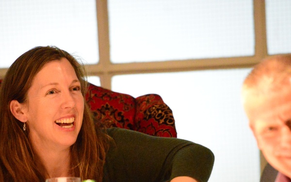 Two adults are seated at a dining table, both smiling and appearing to be engaged in lively conversation. The setting appears to be a formal or semi-formal dinner event, with wine glasses and water bottles visible on the table. The background features a decorative red upholstered chair and a large frosted or backlit window panel.