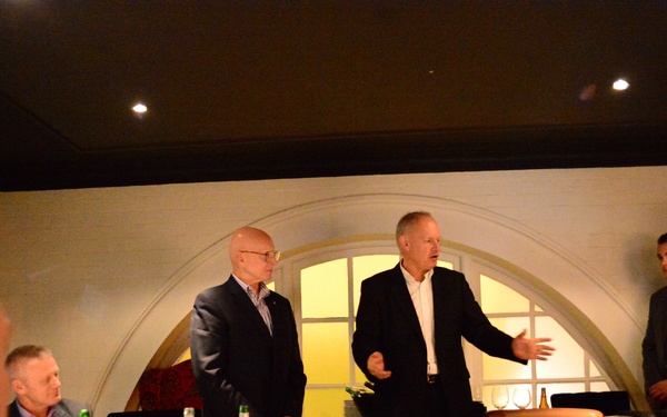 A formal dinner gathering in an upscale restaurant private dining room where two men are standing and addressing seated guests. The setting features an arched window backdrop, white tablecloths, and wine glasses, suggesting a business or social event. One of the standing men appears to be speaking with an open hand gesture while the other stands attentively beside him.
