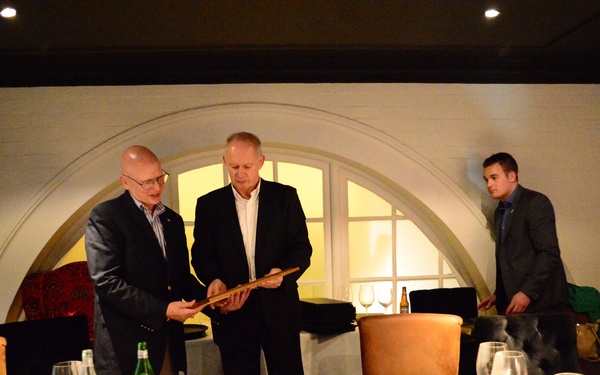 Two men in business attire stand at the head of a dining table in an upscale restaurant setting, appearing to exchange or present a wooden object to one another. The scene suggests a formal dinner event or ceremony, with seated guests observing from around a white-clothed table set with glassware and bottles. A younger man stands in the background near an arched window alcove.