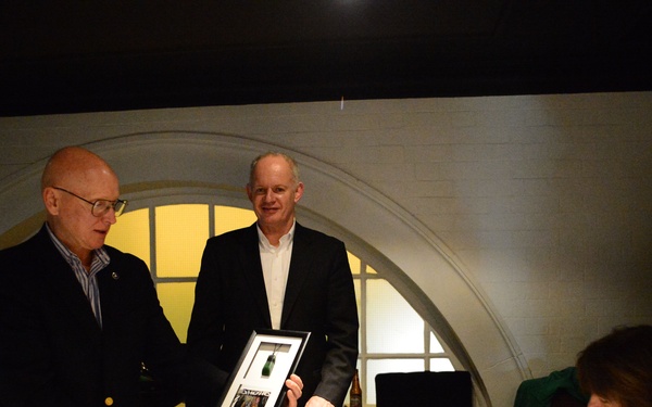 A formal dinner or awards event taking place in an upscale restaurant or venue with arched architectural details. An older bald man wearing glasses is presenting or displaying a framed item, possibly a commemorative plaque or photo collage, while a taller man in a dark suit stands nearby. A woman seated at the table appears to be reviewing documents.