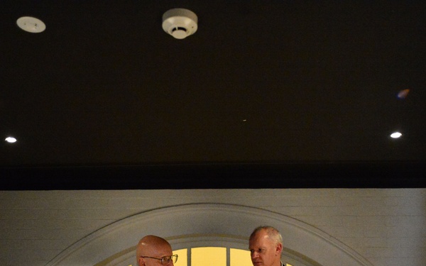 Two men in dark blazers are standing and conversing in what appears to be an upscale restaurant or private dining room. The man on the left, wearing glasses, appears to be speaking while holding something, while the man on the right listens attentively. The setting features a distinctive arched window alcove with warm lighting behind them.