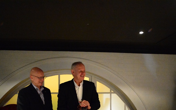 Two men are standing at the head of a dining table in what appears to be a private dining room or restaurant, with one man holding a small award or gift box. The setting suggests a formal dinner event or celebration, with guests seated around a table set with wine glasses, bottles, and tableware. An arched window feature serves as a backdrop behind the standing men.