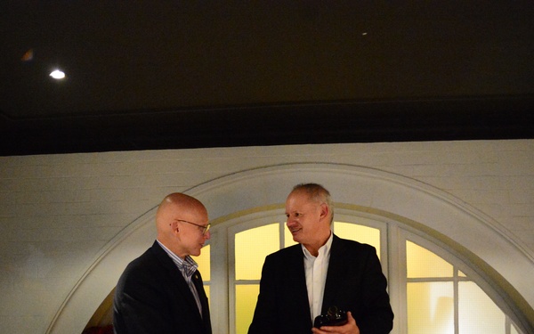 Two men in dark blazers are shaking hands in what appears to be a restaurant or private dining venue, with one man holding a small dark object, possibly a gift or award. A third man is seated to the left, smiling and observing the exchange. The setting suggests a formal or semi-formal dinner event.