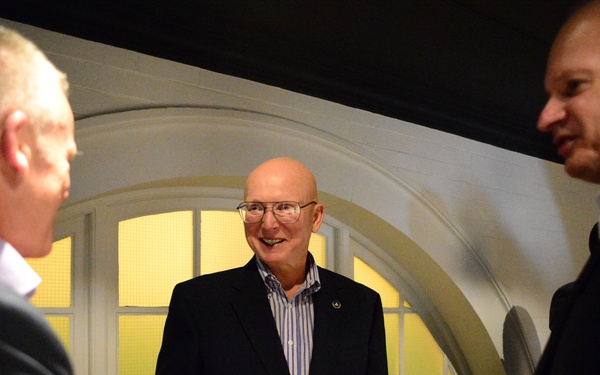 Three men in business attire are engaged in conversation in what appears to be an indoor venue with arched windows. The central figure, a bald man wearing glasses and a dark blazer with gold buttons, is smiling and appears to be the focal point of the interaction. The setting suggests a formal or semi-formal social gathering.