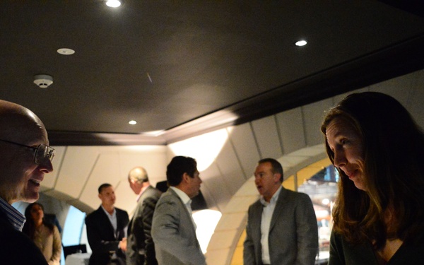 An indoor networking or business event taking place in a dimly lit, modern venue with curved architectural elements. A bald man wearing glasses is shaking hands with a woman in a dark dress in the foreground. Several other attendees are visible in the background engaged in conversation.
