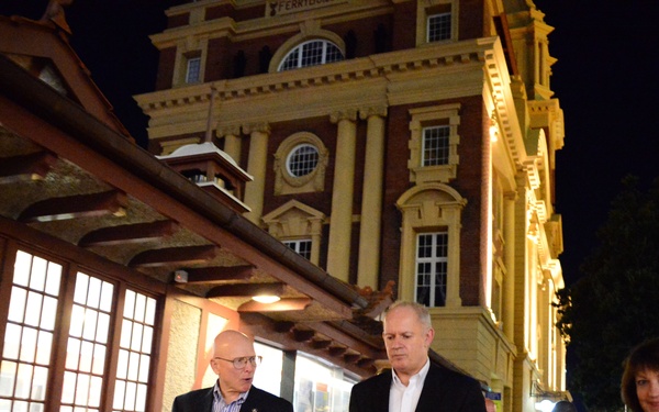 A group of adults walking along a city sidewalk at night in front of an illuminated historic brick building with classical architectural features. Three people are prominently visible in the foreground, with two additional individuals visible in the background. The scene appears to be in a downtown urban area, possibly in Australia or New Zealand based on the architectural style.