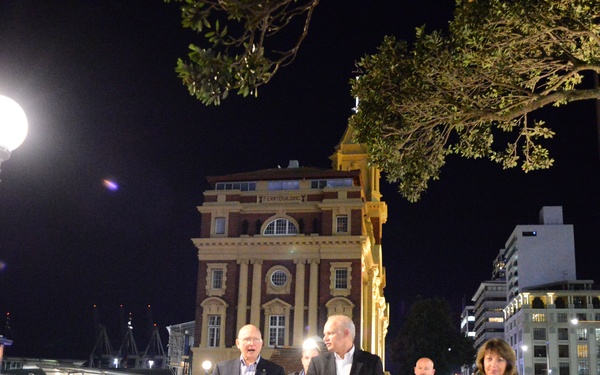 A group of people are walking along a waterfront promenade at night in what appears to be a modern city. The scene is illuminated by a decorative globe street lamp and surrounding urban lighting. The architectural backdrop includes a prominent heritage brick building and contemporary office towers.