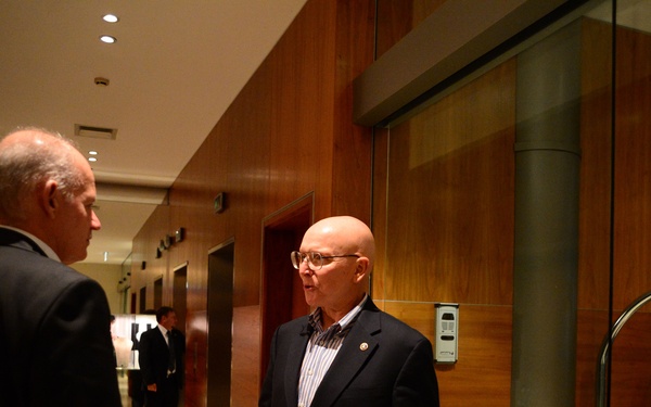 Two men in dark blazers are engaged in conversation in what appears to be a hotel corridor near elevator banks. The man facing the camera is bald, wearing glasses and a navy blazer with a lapel pin, and appears to be speaking with animated hand gestures. The setting is a modern, upscale interior with warm wood-paneled walls and recessed lighting.