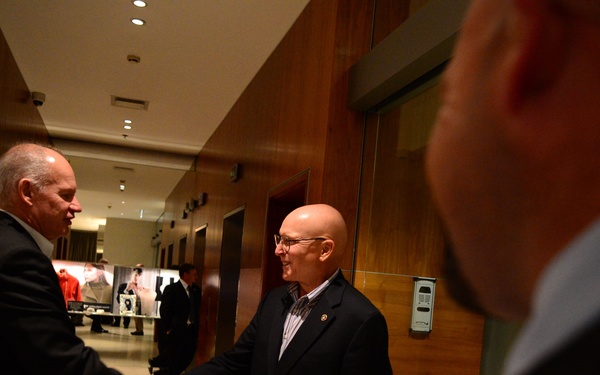 Two men in dark business suits are shaking hands in what appears to be a hotel or upscale building corridor. A third person is partially visible in the foreground on the right side. The setting suggests a formal or professional meeting or greeting.
