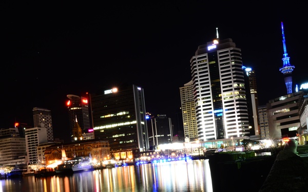 A nighttime cityscape photograph taken from a waterfront vantage point, showing a modern urban skyline with illuminated skyscrapers reflected in calm harbor water. A distinctive tall tower with blue lighting is visible in the background, consistent with Auckland's Sky Tower in New Zealand. The foreground features a stone or concrete seawall, with colorful light reflections — red, yellow, blue, and green — shimmering across the water.
