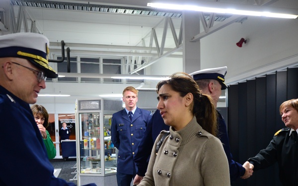 A formal greeting scene inside what appears to be a military or coast guard facility, where a senior uniformed officer is shaking hands with a civilian woman in a tan coat. Multiple uniformed personnel are present in the background, suggesting an official visit or reception event. The setting appears to be an indoor lobby or common area with modern lighting and partitioned spaces.
