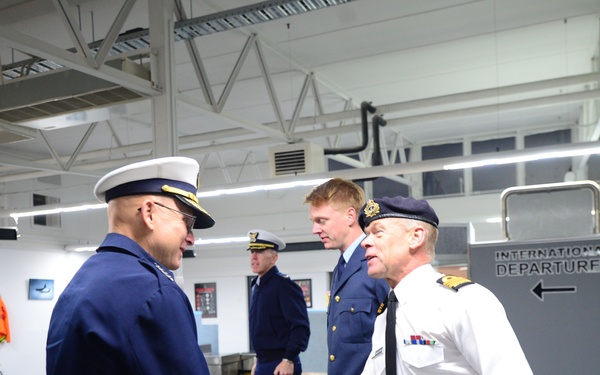 Military or coast guard personnel are exchanging handshakes in what appears to be an airport or maritime facility with an 'International Departures' sign visible in the background. The scene depicts a formal greeting or farewell ceremony between officers in different uniforms, suggesting an inter-agency or international military exchange. At least four uniformed individuals are present in the indoor facility.