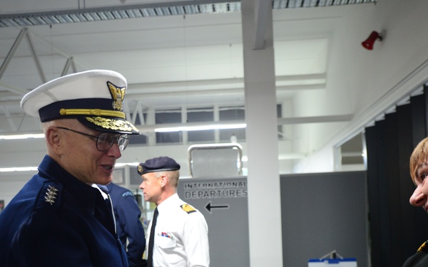 A senior military officer in a dark blue dress uniform with a white peaked cap adorned with gold insignia is shaking hands with another uniformed individual in a dark jacket. The setting appears to be an international departures terminal or customs area of an airport or port facility. Additional uniformed personnel are visible in the background.