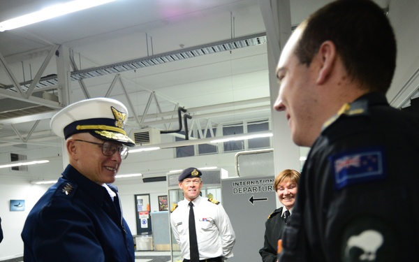 A formal military greeting is taking place at what appears to be an international airport departure area. A senior officer in a dark blue uniform with a white peaked cap is shaking hands with a person wearing a black uniform bearing a New Zealand flag patch. Several other uniformed personnel are present in the background observing the exchange.