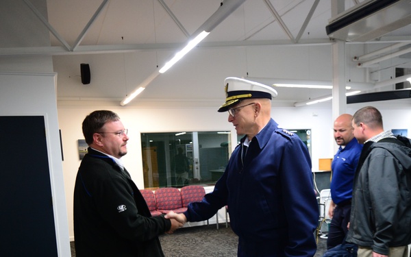 A uniformed military or coast guard officer in a blue dress uniform and white peaked cap is shaking hands with a civilian man in a black jacket inside what appears to be a terminal or institutional building. Two other men are visible in the background, one carrying a large backpack and another holding a black bag. The setting appears to be an official greeting or arrival scenario.