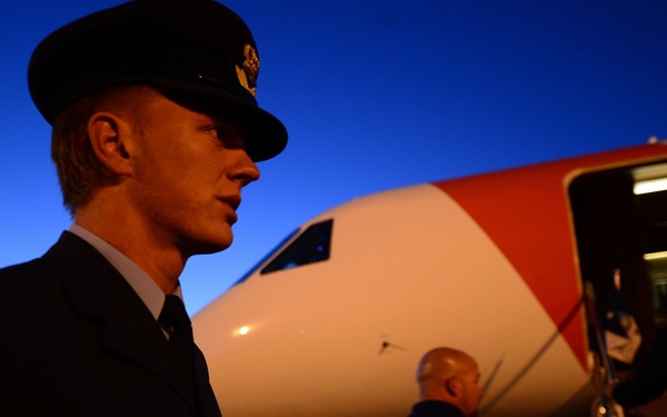 A uniformed military or aviation officer stands on an airport tarmac at dusk or dawn in front of a commercial or government aircraft. The aircraft features a distinctive red, white, and blue livery, and other personnel are visible boarding or standing near the aircraft door. The lighting conditions suggest either early morning or evening operations.