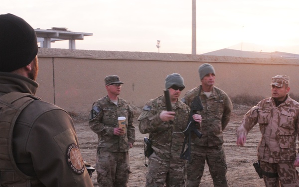 A group of military personnel in mixed camouflage uniforms are gathered outdoors in what appears to be a forward operating base or military compound at dusk or dawn. The soldiers appear to be conducting a briefing or pre-mission coordination, with at least one individual appearing to speak or gesture to the group. Multiple nations appear to be represented based on the different uniform patterns visible.