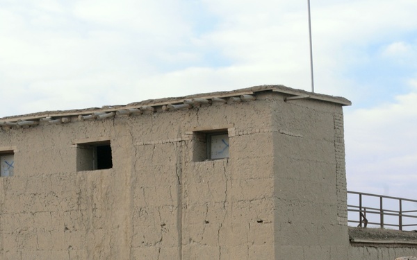 A group of military personnel in mixed camouflage uniforms are gathered outdoors near a mud-brick building, appearing to conduct a weapons training or demonstration exercise. One soldier is actively aiming a rifle while others observe. The setting appears to be a forward operating base or training facility in an arid environment.