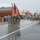 A military parade is taking place on a wet, rain-soaked main street in what appears to be a small American town. Soldiers in Army Combat Uniforms (ACUs) are marching in formation, with a color guard carrying the American flag and what appears to be a unit guidon at the front. Spectators line the sidewalks under umbrellas despite the rainy weather.