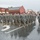 A formation of U.S. Army soldiers in ACU (Army Combat Uniform) camouflage and black berets marches down a wet main street during a rainy parade. The soldiers are marching in disciplined formation along a small-town commercial street lined with brick buildings and spectators. The overcast, rainy conditions have left the road visibly wet with reflections.