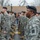 A formation of U.S. Army soldiers in ACU (Army Combat Uniform) digital camouflage pattern are marching in what appears to be a military parade through a small town. The soldiers are wearing black berets and the foreground soldier displays a 3rd Infantry Division patch on his left shoulder. A Christmas wreath visible on a building in the background suggests this is a holiday-season event.