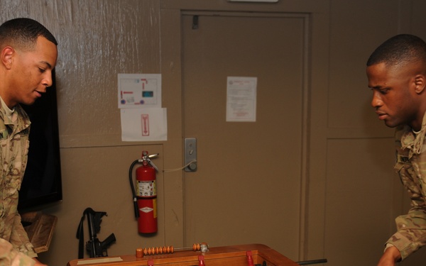 Two U.S. Army soldiers in Operational Camouflage Pattern (OCP) uniforms are playing foosball in what appears to be a recreational room or common area on a military installation. The soldier on the left displays a 1st Cavalry Division patch on his left shoulder. The setting suggests a morale, welfare, and recreation (MWR) facility.