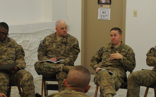 A group of military personnel in US Army MultiCam/OCP camouflage uniforms are seated in a circular arrangement in a plain indoor room, engaged in what appears to be a briefing or discussion session. One central figure appears to be speaking while others listen, some holding notebooks or clipboards. The setting suggests a formal but informal meeting environment, possibly at a military installation.
